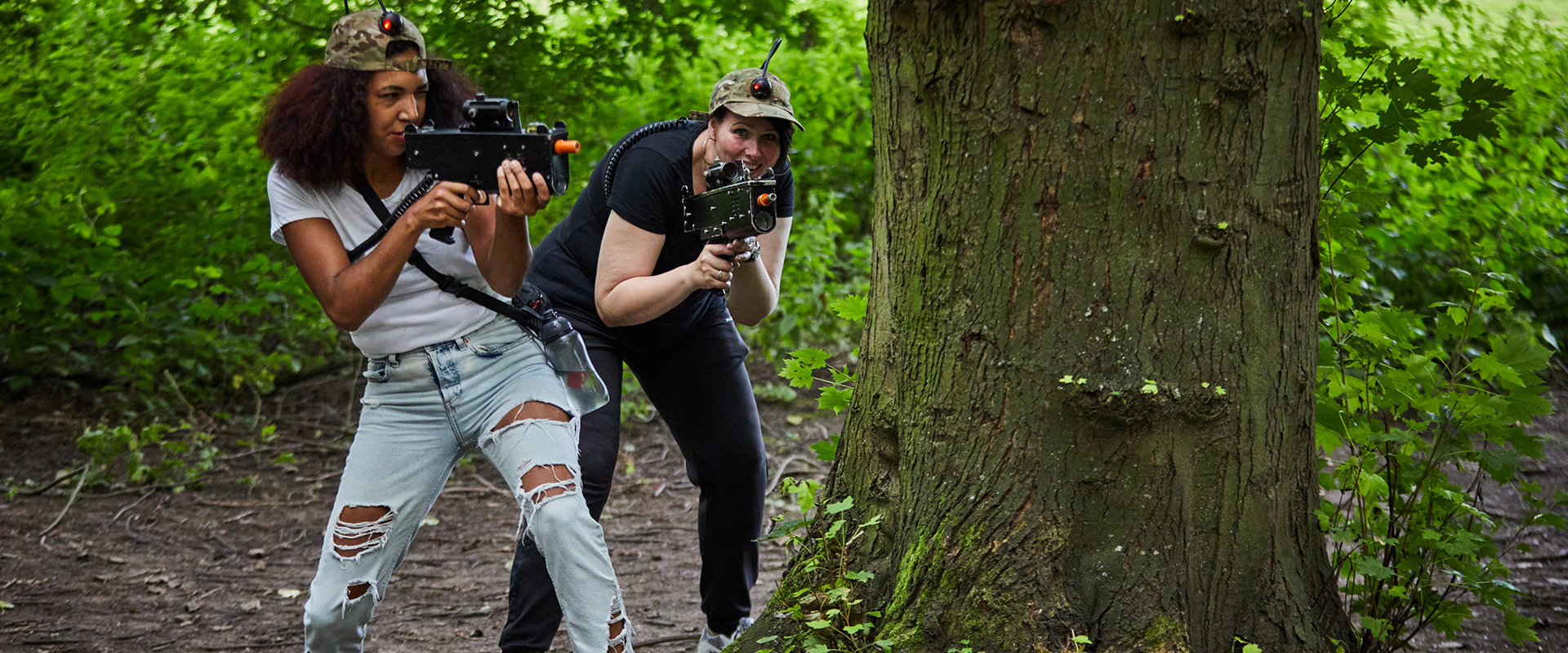 Twee dames aan het lasergamen in het bos. Ze verstoppen zich achter een boom.
